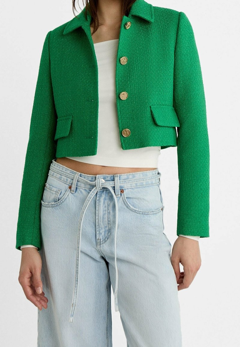 Kobieta ubrana w przykrótką, zieloną kurtkę o fakturze z złotymi guzikami, białą bluzkę crop top oraz jasnoniebieskie dżinsy z wysokim stanem, związane paskiem typu sznurek.