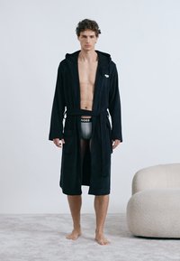 Robe preta com capuz e cinto de tecido texturizado. Modelo usa roupa interior cinza, em pé sobre um tapete claro ao lado de uma cadeira curva.