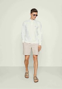 Polo branca de manga comprida, calções bege claro, sandálias castanhas e óculos de sol. Roupa casual com um design e esquema de cores simples.