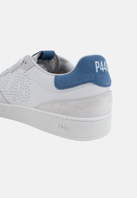Zapatilla blanca con parte superior perforada, acentos de ante gris y una lengüeta de tela azul en el talón. Presenta una suela de goma texturizada con el logo de la marca.