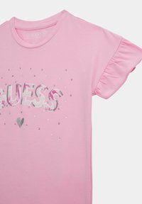 Gaiši rozā T-krekls, izgatavots no organiskās kokvilnas, ar mirdzošiem "GUESS" uzrakstiem un sirdi, kā arī ar viļņainām īsām piedurknēm. Gluda virsma un brīva piegriezuma.