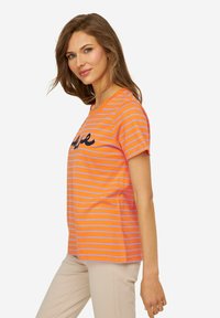 Camiseta de algodón a rayas naranjas con cuello redondo, que presenta rayas moradas y un detalle de texto en cursiva negra en la parte frontal.