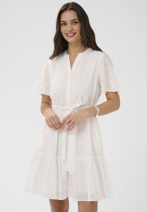 Kobieta w białej, krótkim rękawie sukience z ażurowym wzorem, zapinanej na guziki z przodu, z przewiązanym paskiem w talii, uśmiechająca się z splecionymi rękami.