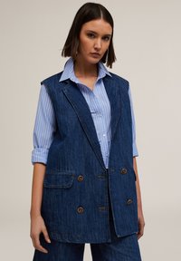 Denimli yelek, çift sıralı düğmeli tasarıma sahip, iki ön cebi, kahverengi düğmeleri ve ham kenarıyla birlikte, açık mavi çizgili bir gömleğin üzerine giyilmiş.