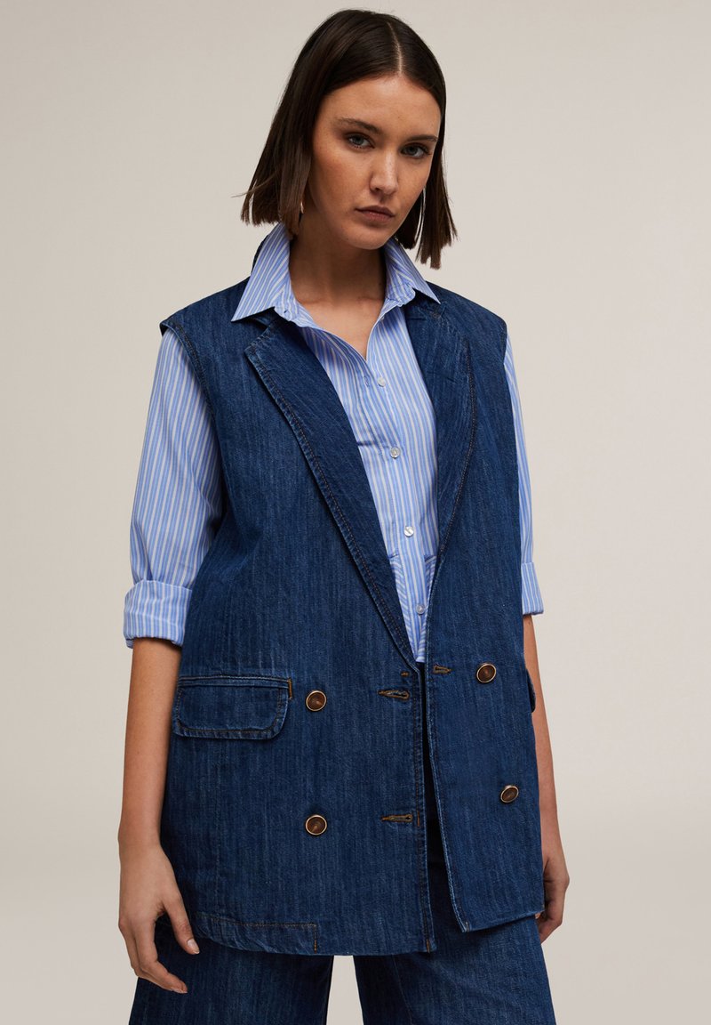 Denimli yelek, çift sıralı düğmeli tasarıma sahip, iki ön cebi, kahverengi düğmeleri ve ham kenarıyla birlikte, açık mavi çizgili bir gömleğin üzerine giyilmiş.