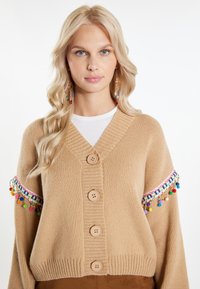 Cardigan lavorato a maglia beige con scollo a V, cinque bottoni e accenti di pompon colorati sulle maniche; tessuto testurizzato con polsini a coste.