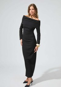Robe noire à épaules dénudées avec des manches longues, coupe slim, tissu texturé et silhouette longue. Associée à des talons noirs et un bracelet en or.