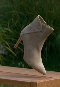 Botine din suede bej, până la glezne, cu vârf ascuțit și toc subțire, având un design fără cusături și coase discrete, expuse pe o suprafață din lemn.