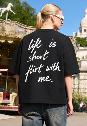 Junge Frau mit blondem Pferdeschwanz, die weiße Sonnenbrille und ein schwarzes T-Shirt mit dem Text "life is short flirt with me" trägt, steht in der Nähe eines Karussells.
