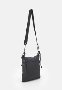 Černá kabelka přes rameno s nastavitelným tkaným popruhem a zipovým vrchem, stojící vzpřímeně na bílém pozadí.