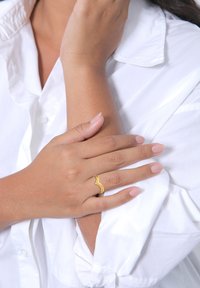 Goldene Wellenring mit Perlenakzenten an einer Hand, die auf einem weißen Hemd ruht. Die Nägel sind in einem sanften, nudefarbenen Ton mit subtiler glänzender Oberfläche lackiert.