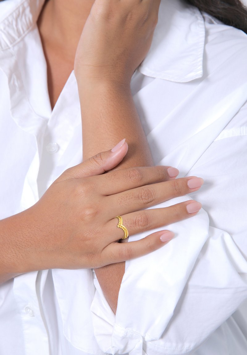 Goldene Wellenring mit Perlenakzenten an einer Hand, die auf einem weißen Hemd ruht. Die Nägel sind in einem sanften, nudefarbenen Ton mit subtiler glänzender Oberfläche lackiert.