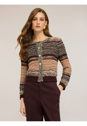 Kvinde med langt, bølget hår iført en flerfarvet, stribet strikket cardigan med knapper og mørke, høj taljede bukser, stående mod en ensfarvet baggrund.