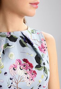 Ljusblå klänning med ett blommönster i rosa, lila och gröna detaljer. Den har en rundad halsringning och subtila veck vid axlarna.