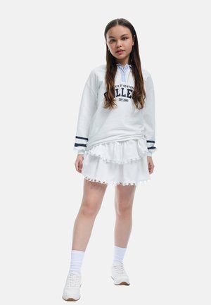 Witte sweatshirt met marineblauwe strepen en grafische print, gecombineerd met een gelaagde witte rok met een scalloped zoom. Witte sneakers en sokken.