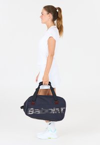 Kvinde i hvidt sportstøj, der holder en sort Babolat sportstaske, stående i profil mod en ensfarvet hvid baggrund.