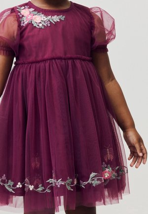 Kind trägt ein lila Tüllkleid mit Puffärmeln und Blumenstickerei an Schulter und Saum, steht vor einem einfarbigen Hintergrund.