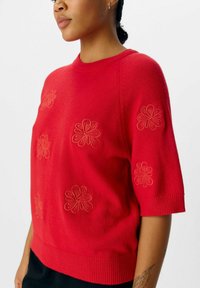 Röd broderad tröja med blommönster, korta ärmar, ribbad nederkant och mjuk textur. Detaljerad sömnad i ljusare röd.