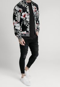 Květinová bomber bunda v černé barvě s červenými a bílými vzory ibišků; doplněná černými potrhanými úzkými džíny a bílými teniskami.