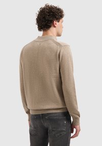 Man met krullend haar, gekleed in een beige trui en donkergrijze jeans, met zijn rug naar de camera gekeerd tegen een effen lichtgrijze achtergrond.