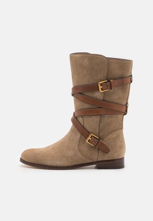 Suede ankle boot in taupe with dark brown leather straps and gold buckles. Flat sole, rounded toe, and smooth texture. Side design detail included.