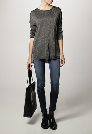 Graues, metallisches Langarmoberteil kombiniert mit dunklen Skinny-Jeans und schwarzen Ankle-Boots. Eine schwarze Tote-Tasche mit einem kreisförmigen Design ist ebenfalls enthalten.