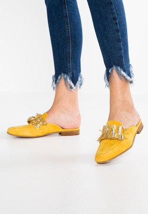 Żółte zamszowe buty wsuwane z frędzlami i metalowymi akcentami, z wąskim noskiem i niskim drewnianym obcasem. Noszone z postrzępionymi niebieskimi dżinsami.