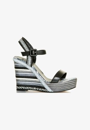 Sandalia cuña negra con una amplia tira en la puntera y tira ajustable en el tobillo. La cuña a rayas presenta texturas y patrones en gris, negro y blanco.