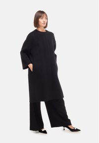 Cappotto oversize nero con scollo rotondo, maniche lunghe e tasche laterali, abbinato a pantaloni neri a gamba larga e tacchi a punta con un accento metallico.