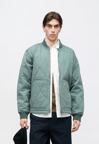 Ανοιχτοπράσινο καπιτονέ μπουφάν με φερμουάρ μπροστά, ριμπ μανσέτες, δύο εμπρόσθιες τσέπες και μικρή λογότυπο διακοσμητικό. Φορεμένο πάνω από λευκό πουκάμισο και καφέ τοπ.