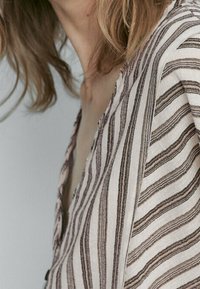 Lekki, w paski top z materiału, który ma naprzemienne ciemno- i jasnobrązowe linie; dekolt w serek, luźny krój i miękka tekstura.