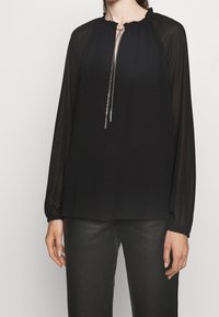 Chemisier noir à manches longues avec manches transparentes, encolure froncée et lien avant en chaîne argentée, associé à un pantalon en cuir noir sur une personne debout.