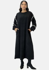 Abito maxi in corduroy nero con maniche lunghe, caratterizzato da bordi bianchi e tasche laterali. Il materiale ha una finitura testurizzata e una vestibilità rilassata.