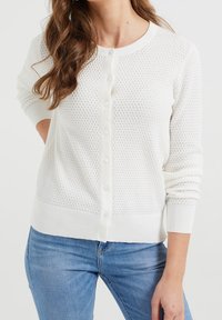 Bílý pletený kardigan s texturovaným vzorem, kulatým výstřihem a zapínáním na knoflíky. Žebrované manžety a lem, nošený přes modré džíny.