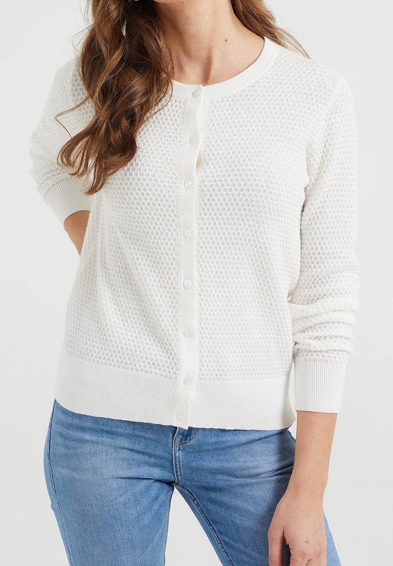 Bílý pletený kardigan s texturovaným vzorem, kulatým výstřihem a zapínáním na knoflíky. Žebrované manžety a lem, nošený přes modré džíny.