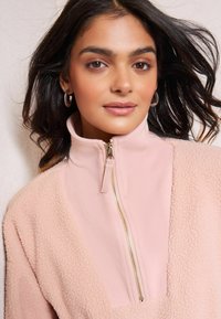 Heled roosa pool-zaamiga pullover ühendab ribatud ja karvase tekstuuri, millel on kuldne tõmblukk ja kõrge krae. Sile kangas kontrastib pehme viimistlusega.