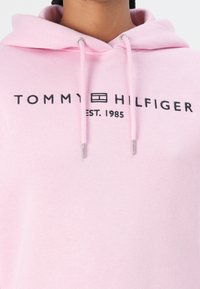 Svetloružová mikina s kapucňou na šnúrku, s čiernym logom "TOMMY HILFIGER" a textom "EST. 1985". Mäkká tkanina s kovovými akcentmi v striebornej farbe.