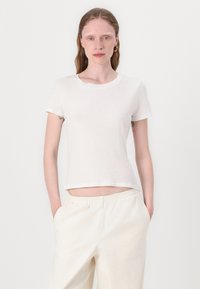 Balta trumpa rankovė t-shirt su apvalia iškirpte, pagaminta iš griovelio tipo medžiagos. Dėvima su šviesios spalvos, elastingais juosmeniu kelnėmis. Fonas neutralus.