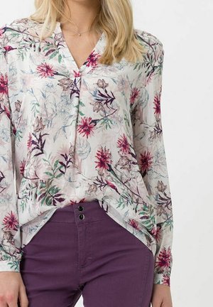 Femme portant un chemisier blanc à motifs floraux roses et verts, rentré dans un pantalon violet taille haute à doubles boutons.