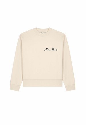 Femme aux cheveux noirs mi-longs, portant des lunettes de soleil écaille de tortue et un sweat crème avec le texte "Mira Paris" sur la poitrine.