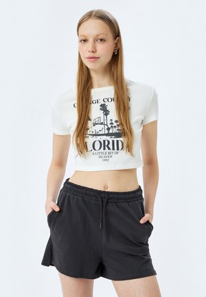Geknipte witte t-shirt met een grafische print van palmbomen en tekst; gecombineerd met losse zwarte shorts met een elastische tailleband en zakken.