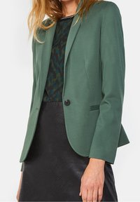 Green tailored blazer with a smooth fabric, single button closure, and slim fit. Features a notch collar and two side pockets.