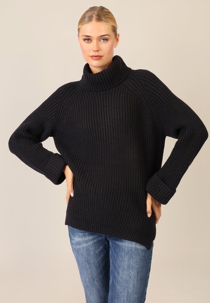 Maglione nero a coste con colletto alto e piegato e spalle scese. Presenta maniche lunghe e una vestibilità ampia. Indossato con jeans blu.