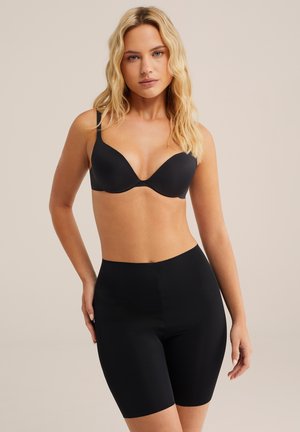 Lyshåret kvinde iført en sort bh og sorte figursiddende shorts med høj talje, stående mod en ensfarvet beige baggrund.