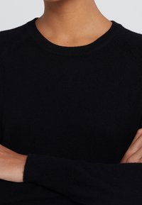 Schwarzer Strickpullover mit rundem Ausschnitt und langen Raglanärmel. Die Textur ist weich und gerippt, was eine enge Passform betont.