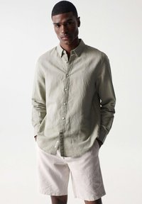 Camisa de botón verde oliva claro hecha de tela texturizada, combinada con pantalones cortos beige claro. La camisa tiene un cuello clásico y un bolsillo de parche.