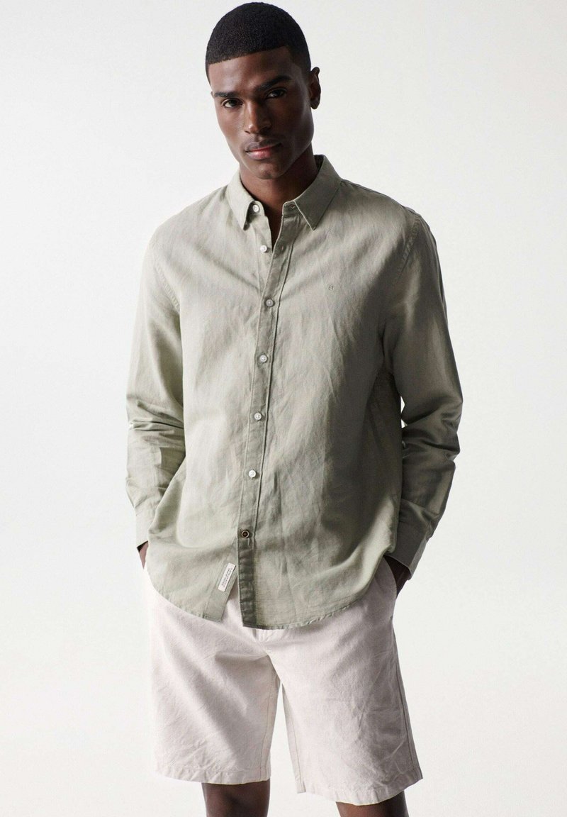 Camisa de botón verde oliva claro hecha de tela texturizada, combinada con pantalones cortos beige claro. La camisa tiene un cuello clásico y un bolsillo de parche.