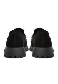 Černé semišové boty s tlustou, strukturovanou černou gumovou podrážkou. Zaoblený tvar s nízkým střihem. Pohled zezadu ukazuje švy a minimalistický design.