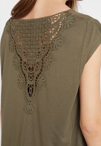 Top verde oliva con intrincados detalles de encaje en la parte posterior, con un patrón floral y un diseño con corte, hecho de una tela suave.