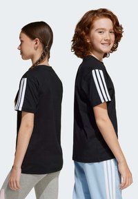 T-shirt en coton noir à manches courtes, avec trois rayures blanches sur l'épaule. Porté par deux jeunes mannequins, l'un ayant une tresse.